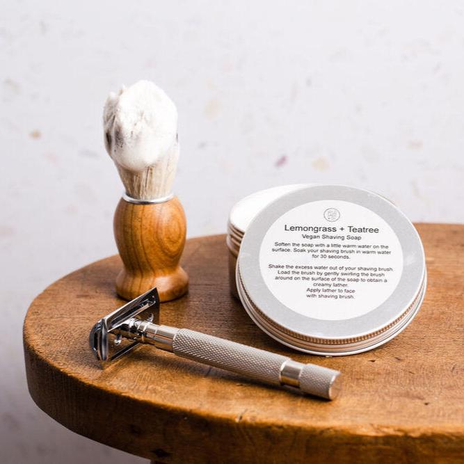 Wooden shaving brush on table next to metal razor and lemongrass & teatree shave soap tin