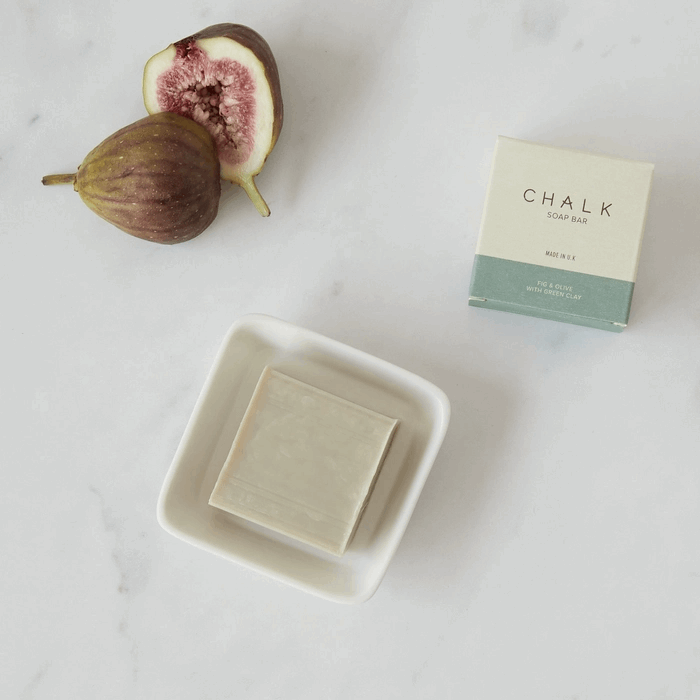 White porcelain square soap dish from chalk with fig soap. Sat on marble vanity counter top, next to a cut fig.