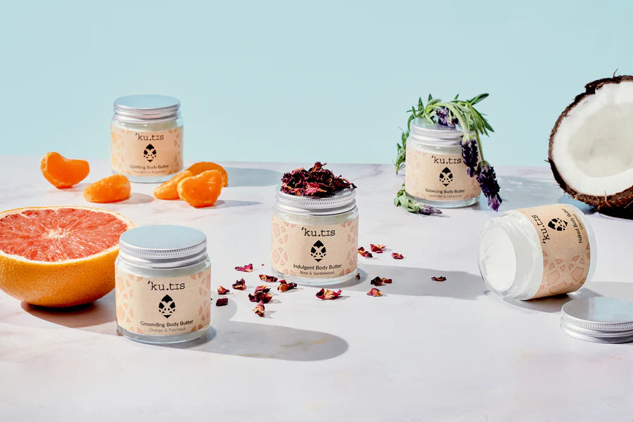 Kutis body balms displayed on a table next to a coconut, mandarin slices, a cut grapefruit, a sprig of lavender and scattered rose petals. Jars are open and on their side.